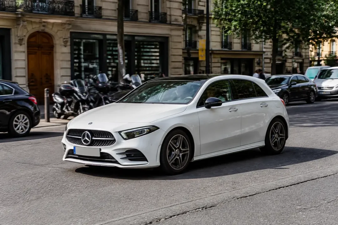 Voiture Mercedes Classe A blanche circulant en ville sur une route bordée d'arbres