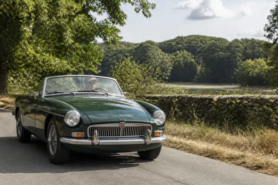Voiture ancienne MG verte sur route de campagne bordée d'arbres et mur pierre